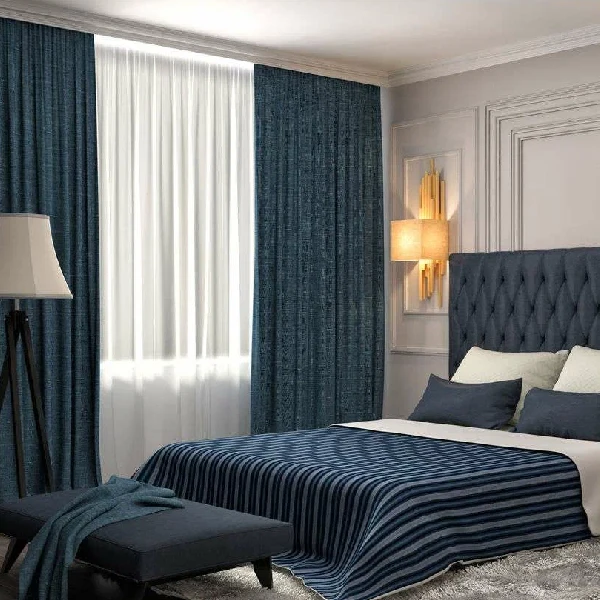 A bedroom with blue and white striped bedding, a tufted blue headboard, and a sheer white curtain flanked by dark blue curtains.